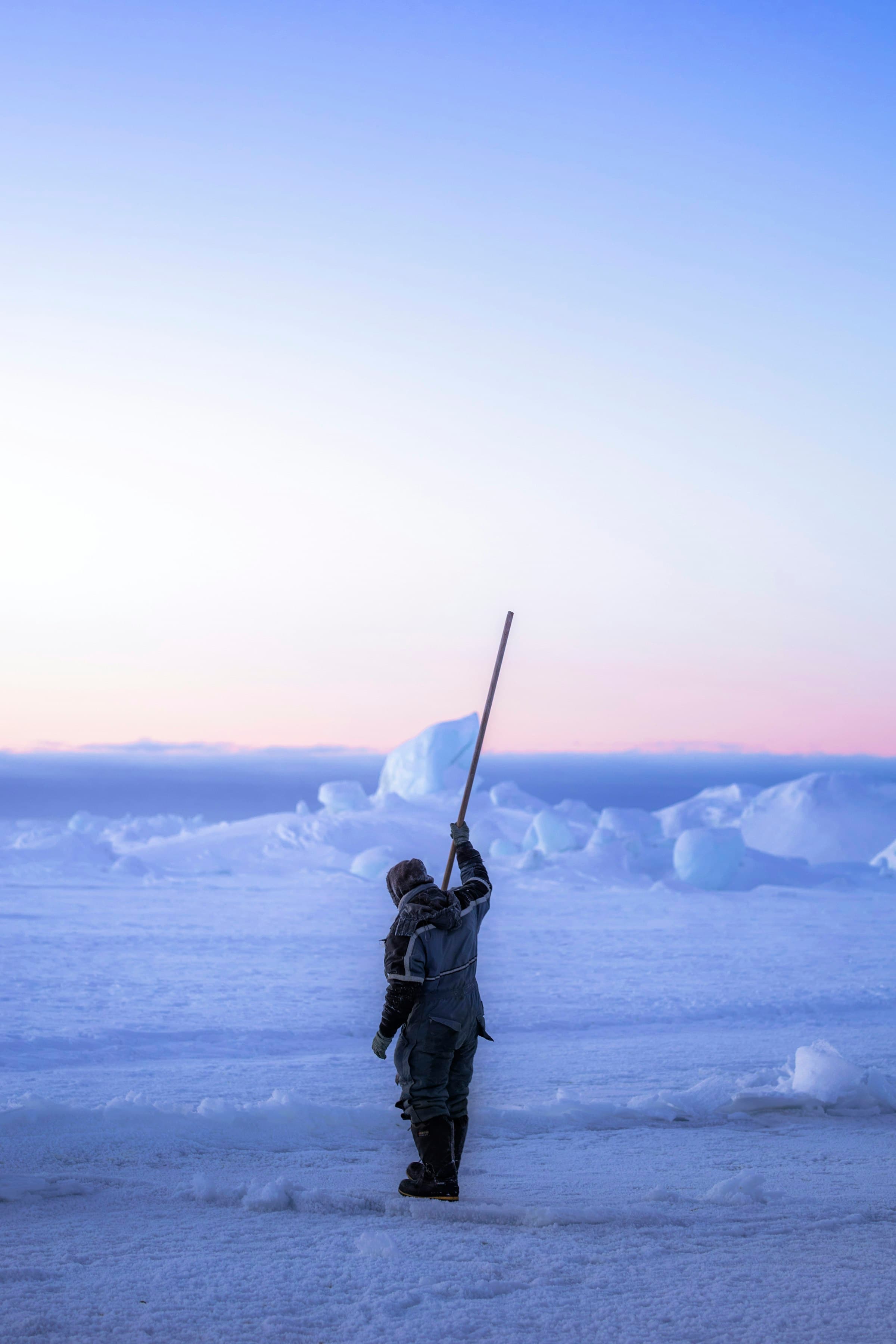 A figure on sea ice raising a long pole towards the horizon